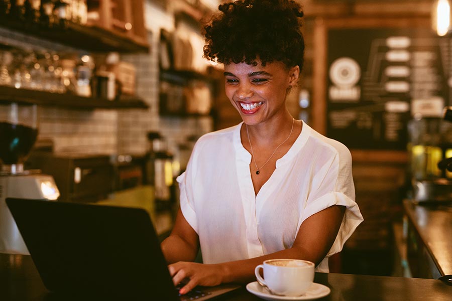 Client Center - Young Woman Grinning as She Uses a Laptop in a Dimly Lit Cafe, a Cappuccino on Her Table